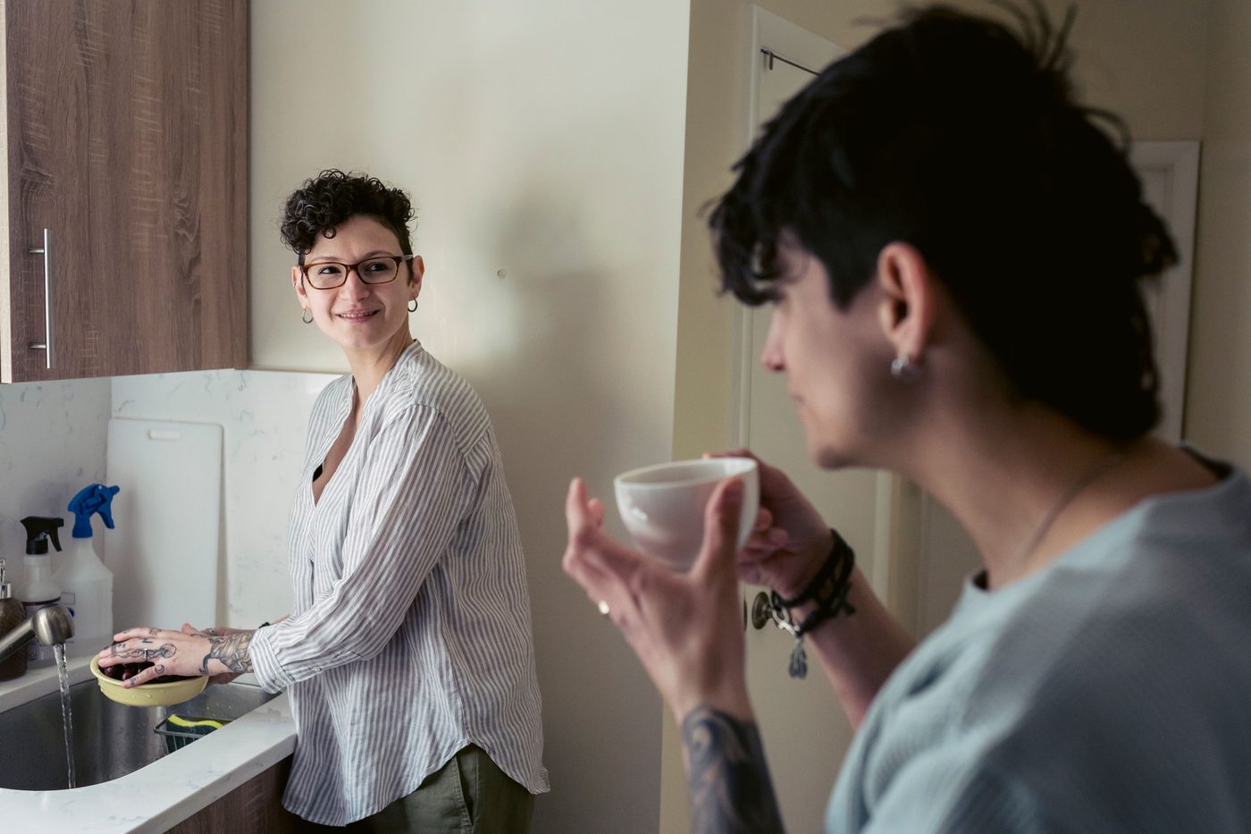 Two housemates talking in their kitchen, one washing cherries at the sink while the other holds a cup of coffee